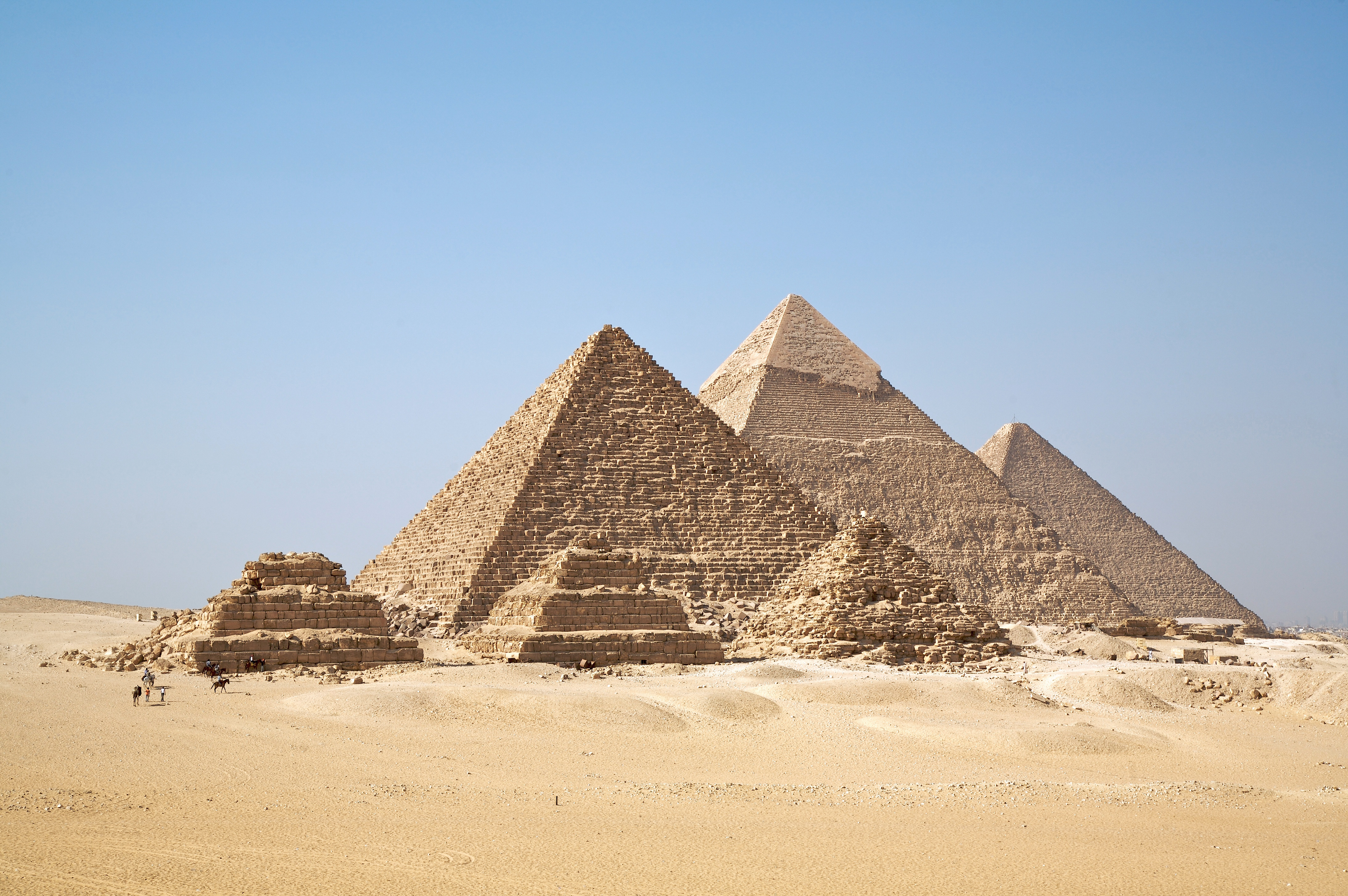 The Pyramids of Giza rise in the sand desert under a clear blue sky, with several small visitors visible in the foreground.