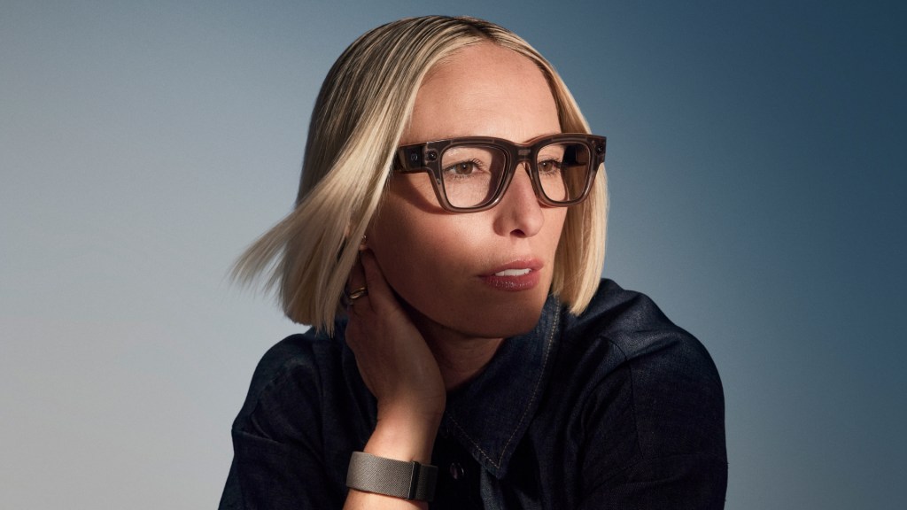 A person with blonde hair and dark-framed glasses wearing a dark blue jacket against a blue-grey background.