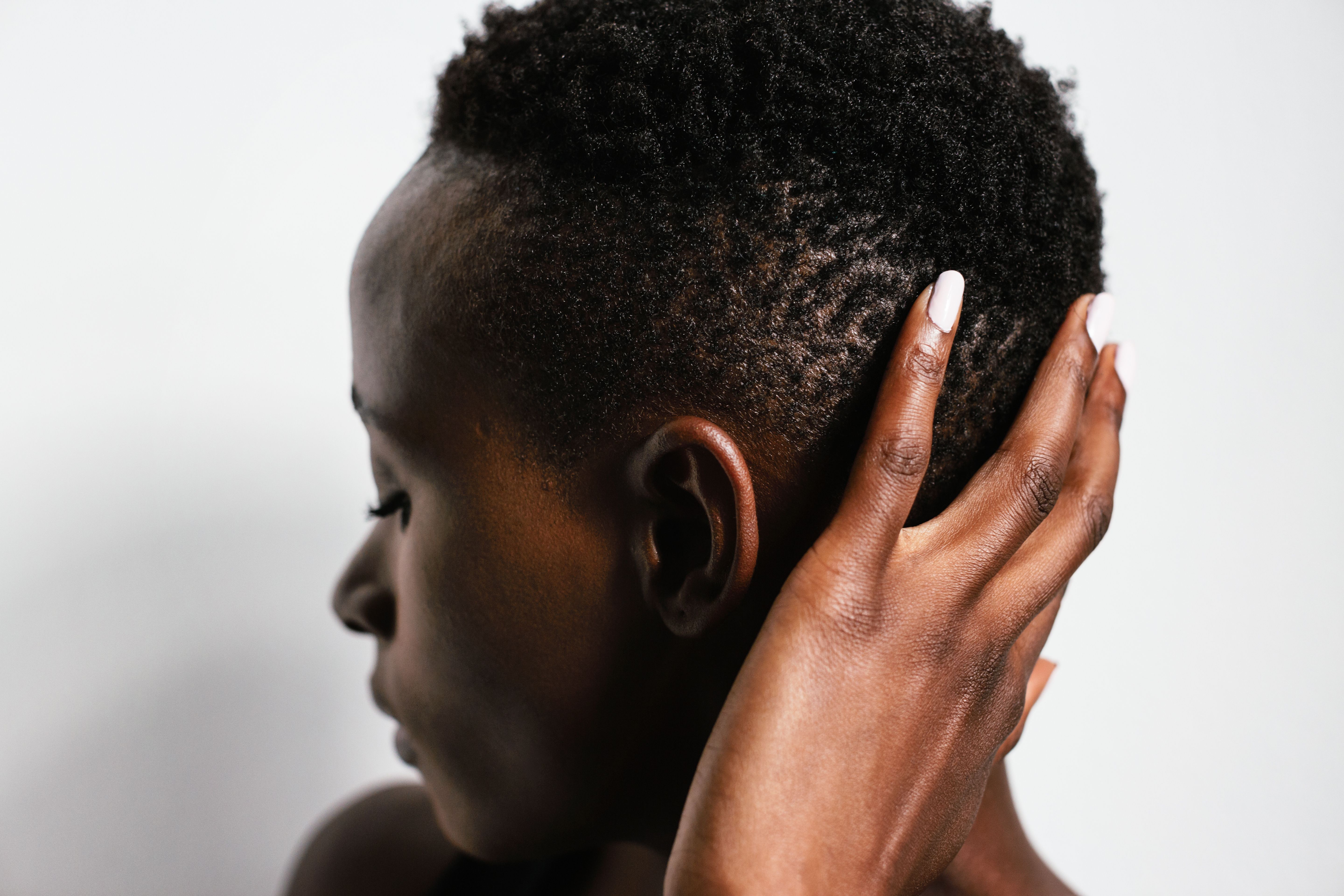 A person in profile touches their curly natural hair near the scalp with their hand, showing the top of the head and ear area.