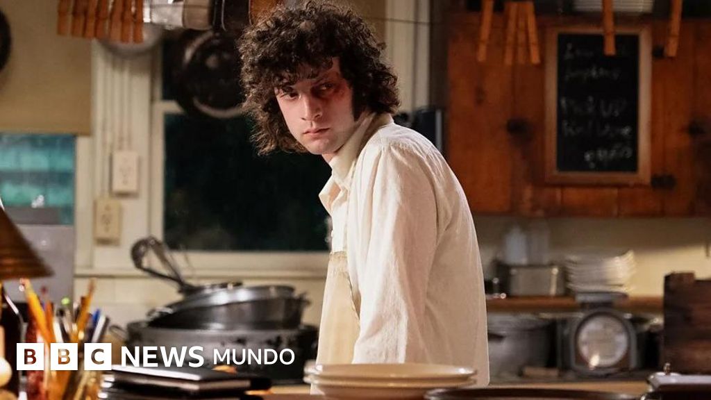 A young man with dark curly hair wearing a light white shirt stands in a kitchen, looking directly at the camera with a serious expression.