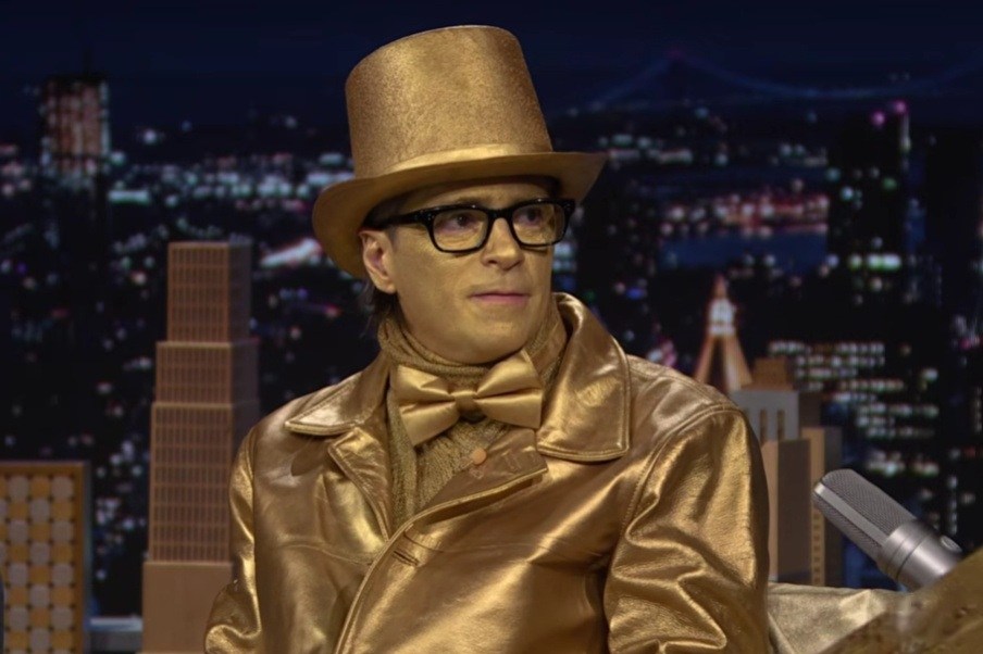 A man wearing glasses dressed entirely in gold, including a golden top hat, shining jacket and bow tie, sitting in a television studio with a city skyline lit up at night behind him.