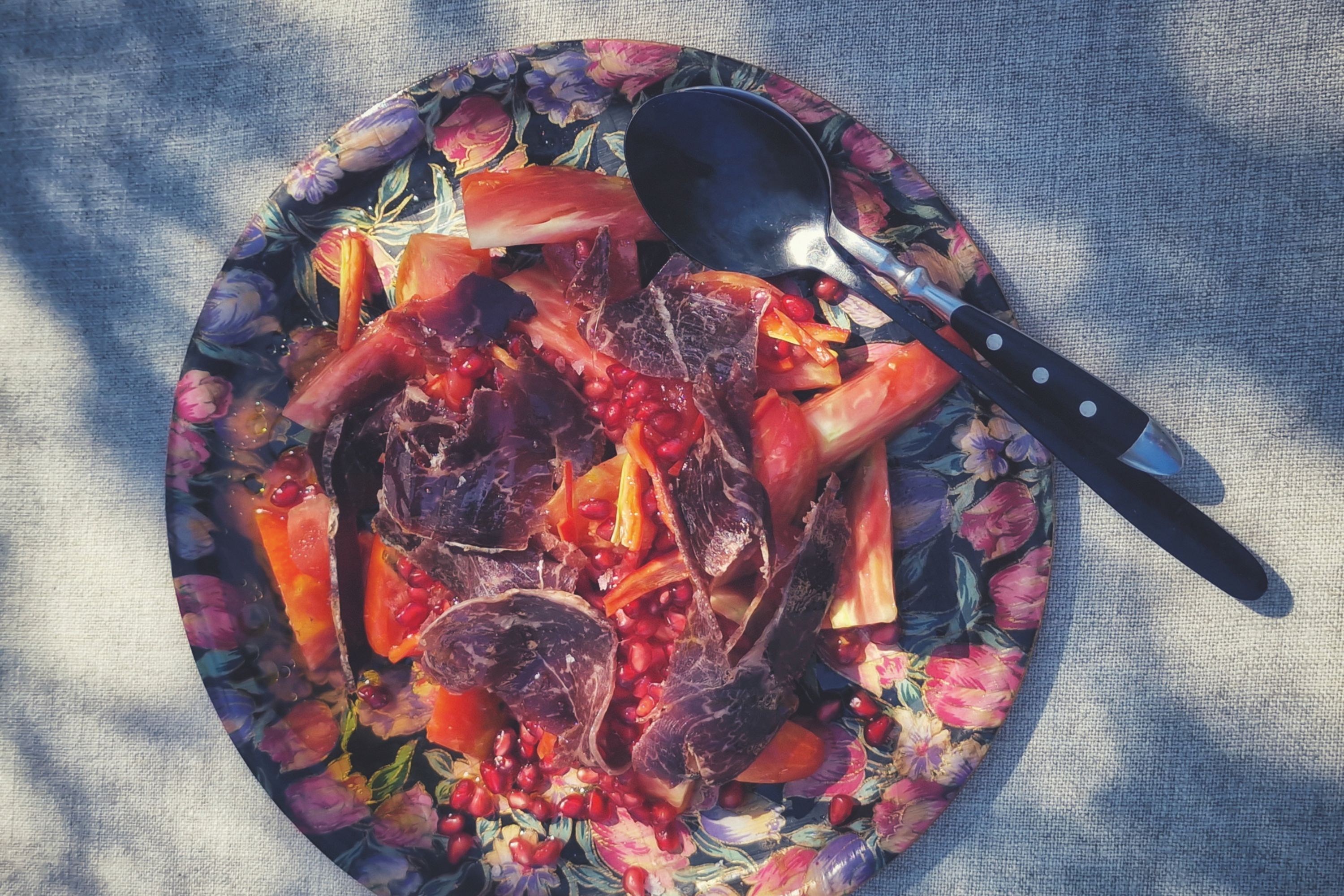 Un plato decorativo con estampado floral de colores vibrantes contiene carnes rojas crudas junto a pimientos y una cuchara metálica negra.