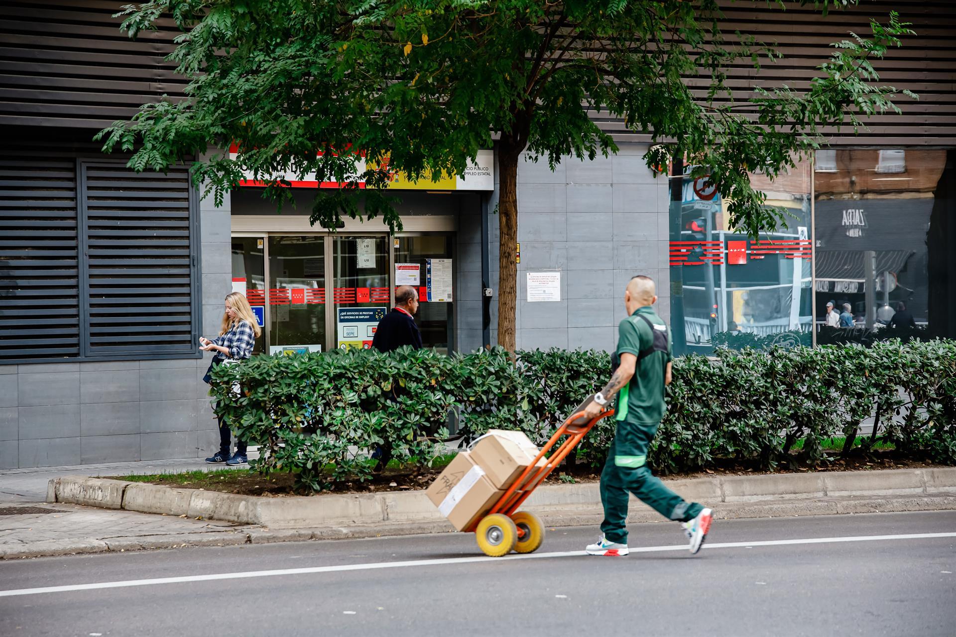 Un repartidor con uniforme verde transporta una caja en un carrito de mano por una calle urbana frente a una tienda con decoraciones rojas.