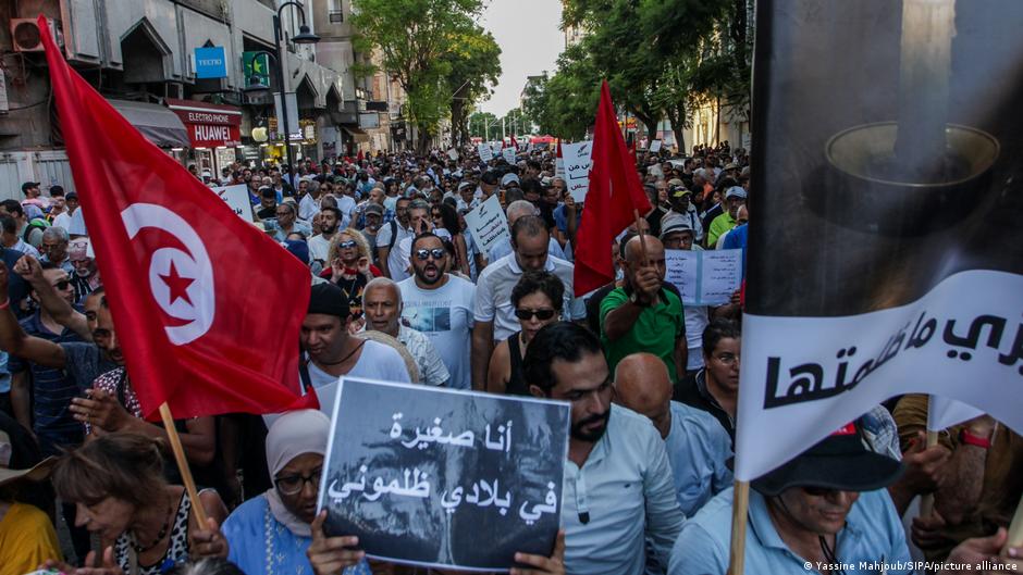 Una multitud de manifestantes llena una calle de la ciudad portando banderas tunecinas rojas y blancas, pancartas escritas en árabe, y carteles con mensajes de protesta.