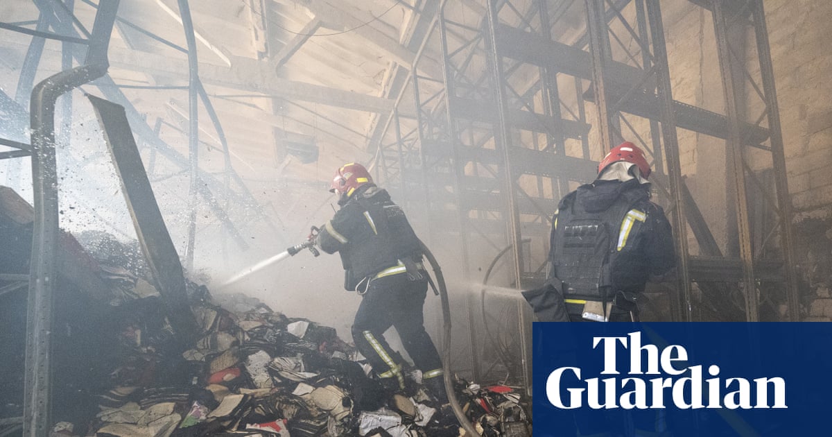 Firefighters in protective gear spray water on smoldering ruins of a heavily damaged building filled with metal structural beams and debris.