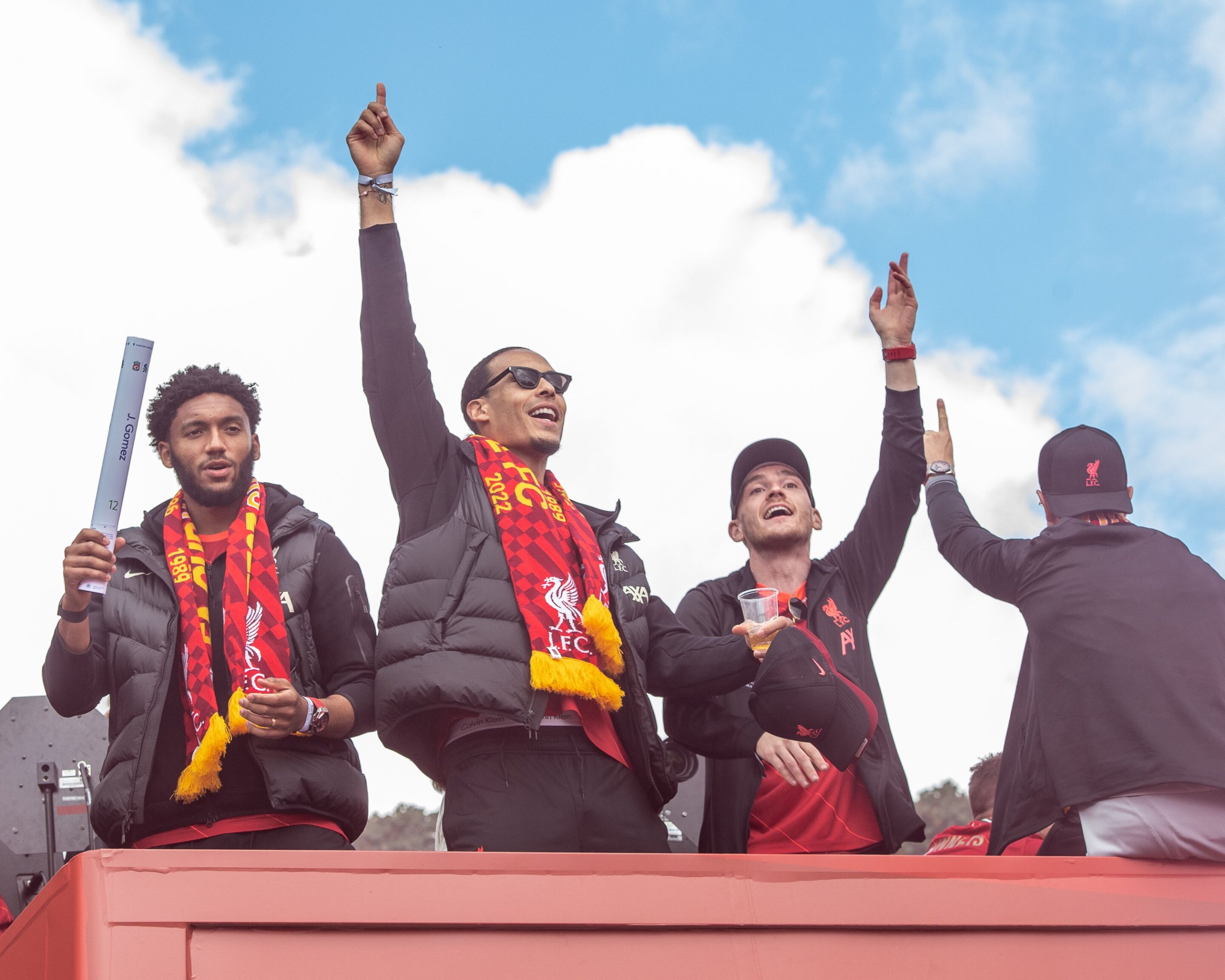 Four men celebrating on top of a red open-top bus, with arms raised to the sky, wearing scarves and accessories of an English football team, under a blue sky with white clouds.
