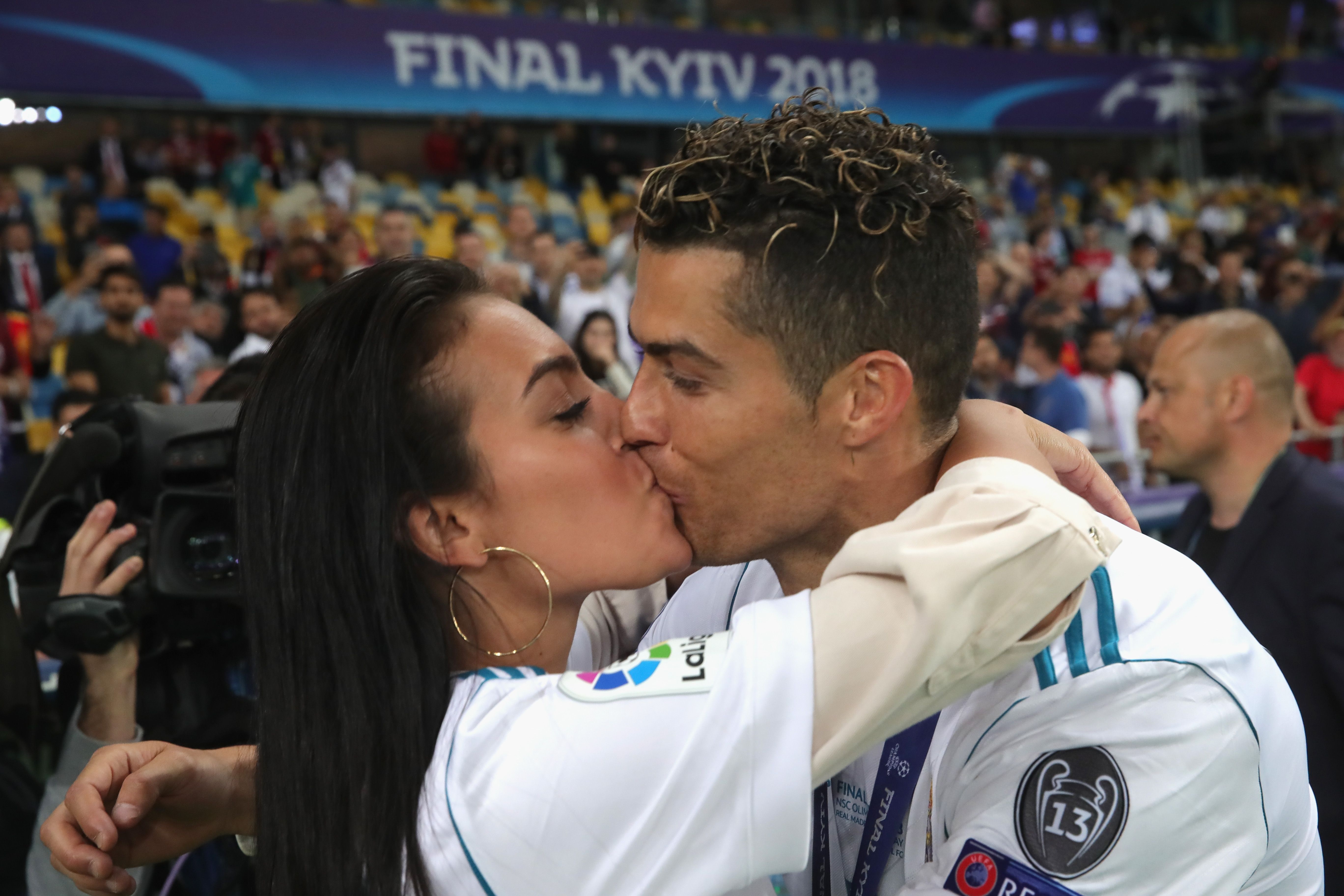 A man and woman kiss at a sports stadium with crowds visible in the background.