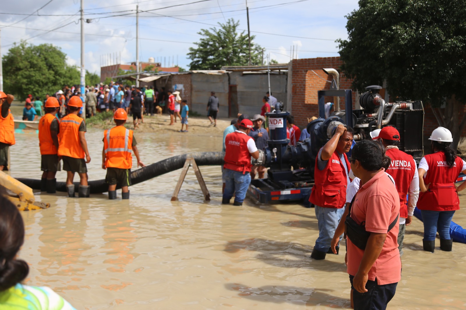 Workers and volunteers in orange and red safety vests respond to flooding on a waterlogged street in a residential community, with brick houses in the background and drainage equipment visible.