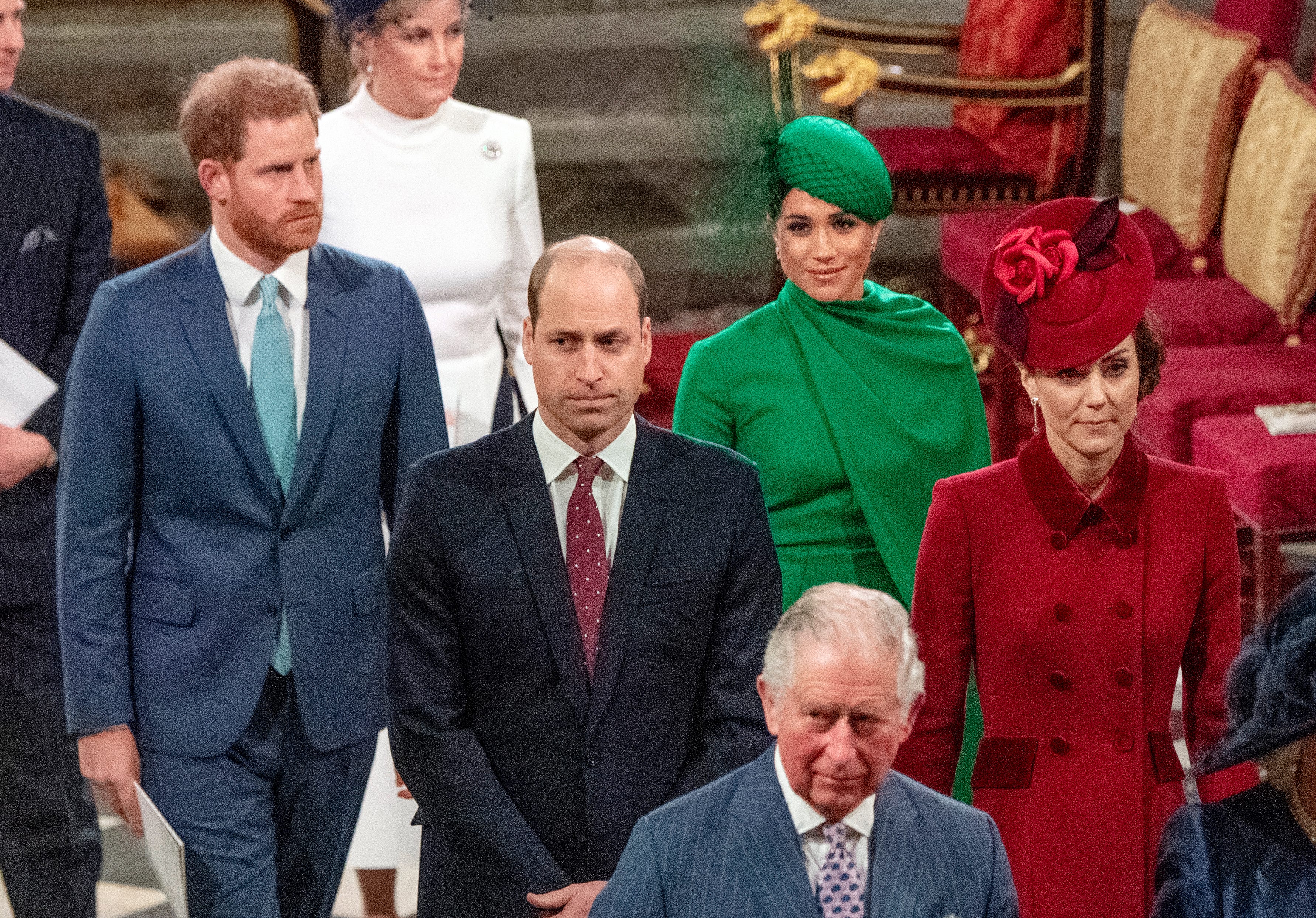 Members of the British Royal Family stand together in formal attire at an official indoor ceremony, with some wearing decorative hats and formal clothing in navy, white, green, and red.