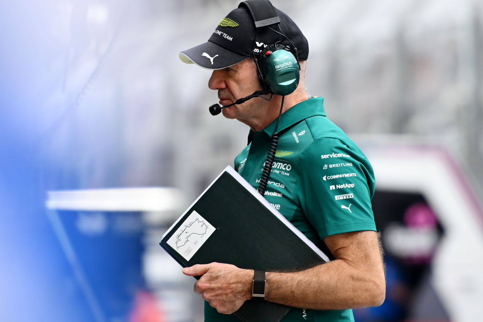 Un ingeniero de Aston Martin viste un uniforme verde con auriculares y sostiene una tableta con un diagrama de circuito mientras observa a un lado en lo que parece ser una sesión de entrenamiento o carrera.