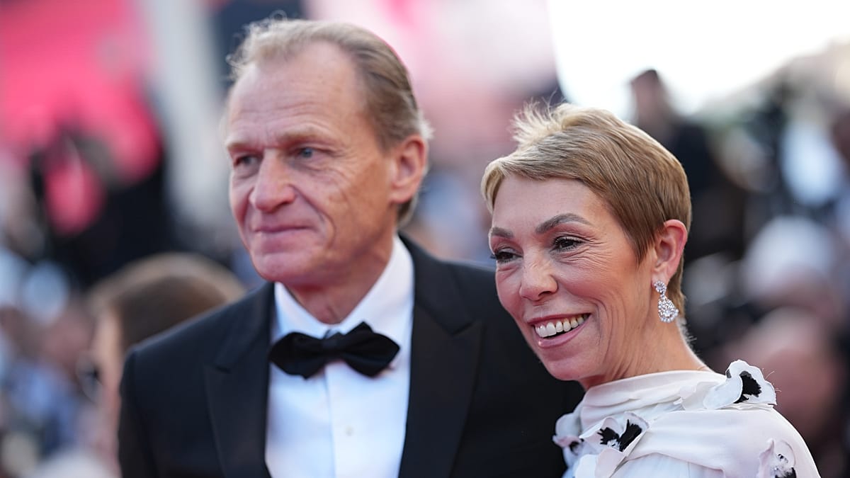An elderly man in a tuxedo with a black bow tie and a woman with short blonde hair, diamond earrings and a white dress with black details smile at a formal event.