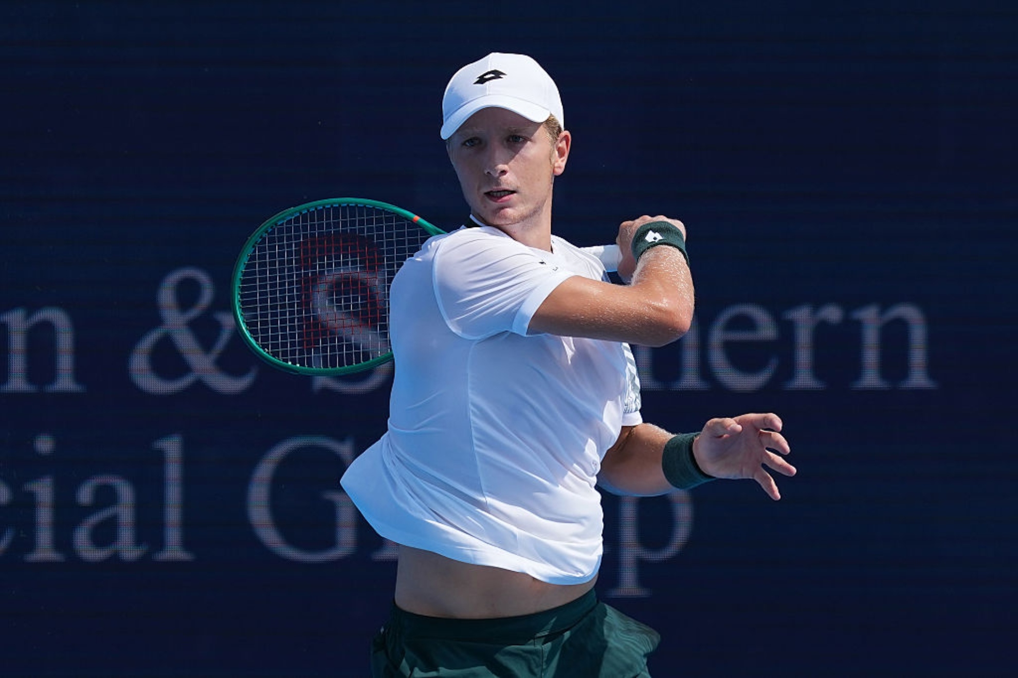 A tennis player dressed in white with a white cap holds a tennis racket with a green frame while in playing position on the court.