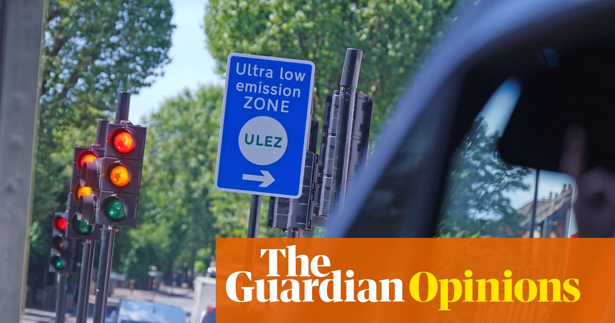 A blue ULEZ (Ultra Low Emission Zone) sign with a white arrow pointing right, mounted on a street pole next to traffic lights showing red and amber, with green trees lining a London street in the background.