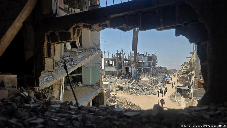 Vista desde el interior de un edificio destruido mostrando un paisaje urbano devastado con ruinas de construcciones, escombros esparcidos y personas caminando entre los edificios dañados bajo un cielo despejado.