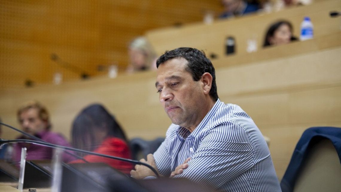 A middle-aged man in a blue and white striped shirt speaks at a microphone during a legislative session in a hemicycle.