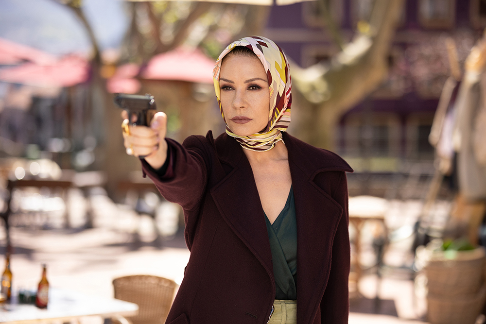 A woman with a yellow and white headscarf wrapped around her head points a gun at the camera, wearing a dark brown blazer, in a setting that appears to be an Arab courtyard or square.