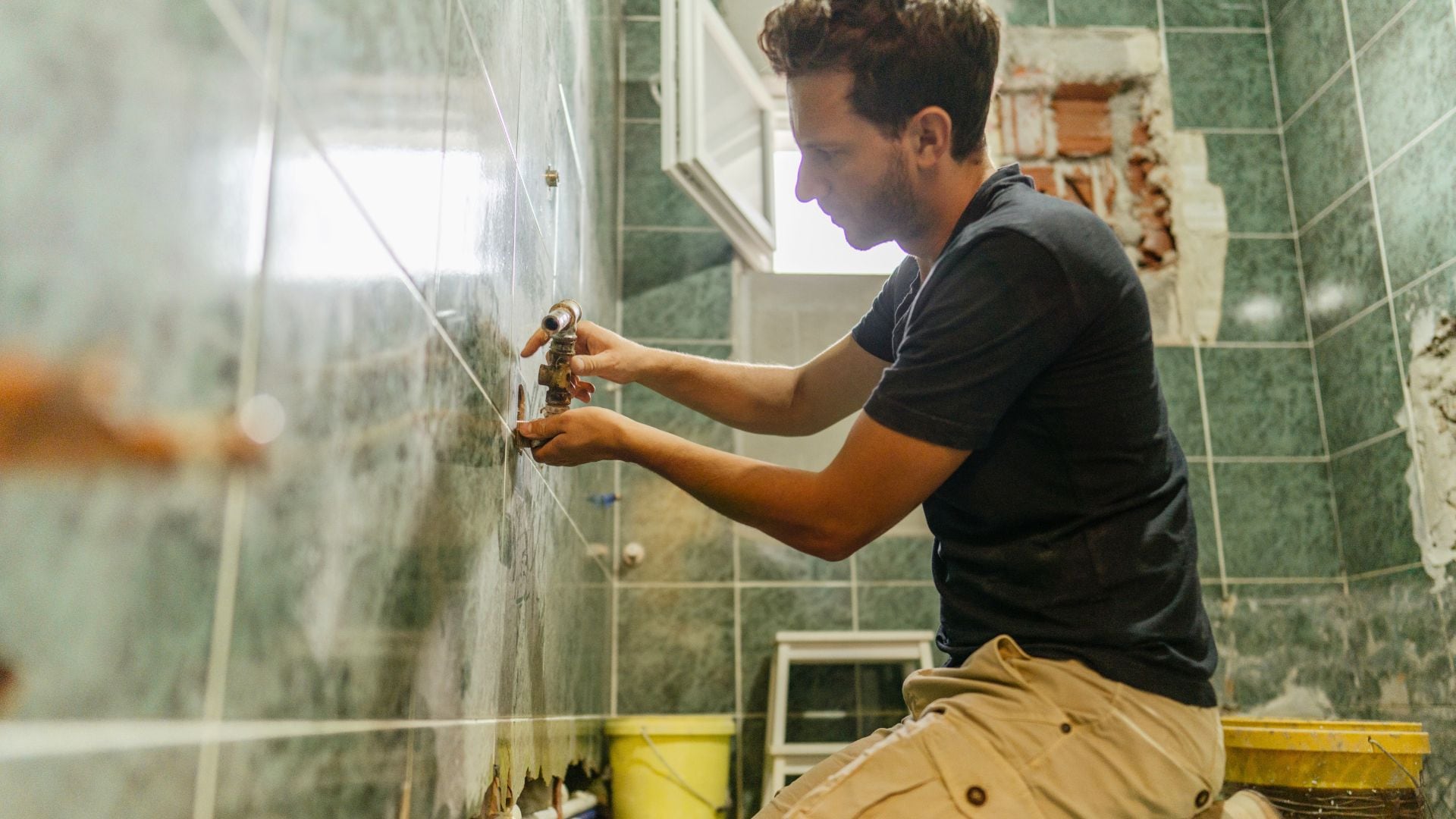 A man in a black shirt installs a water valve on a green tile wall inside a bathroom.