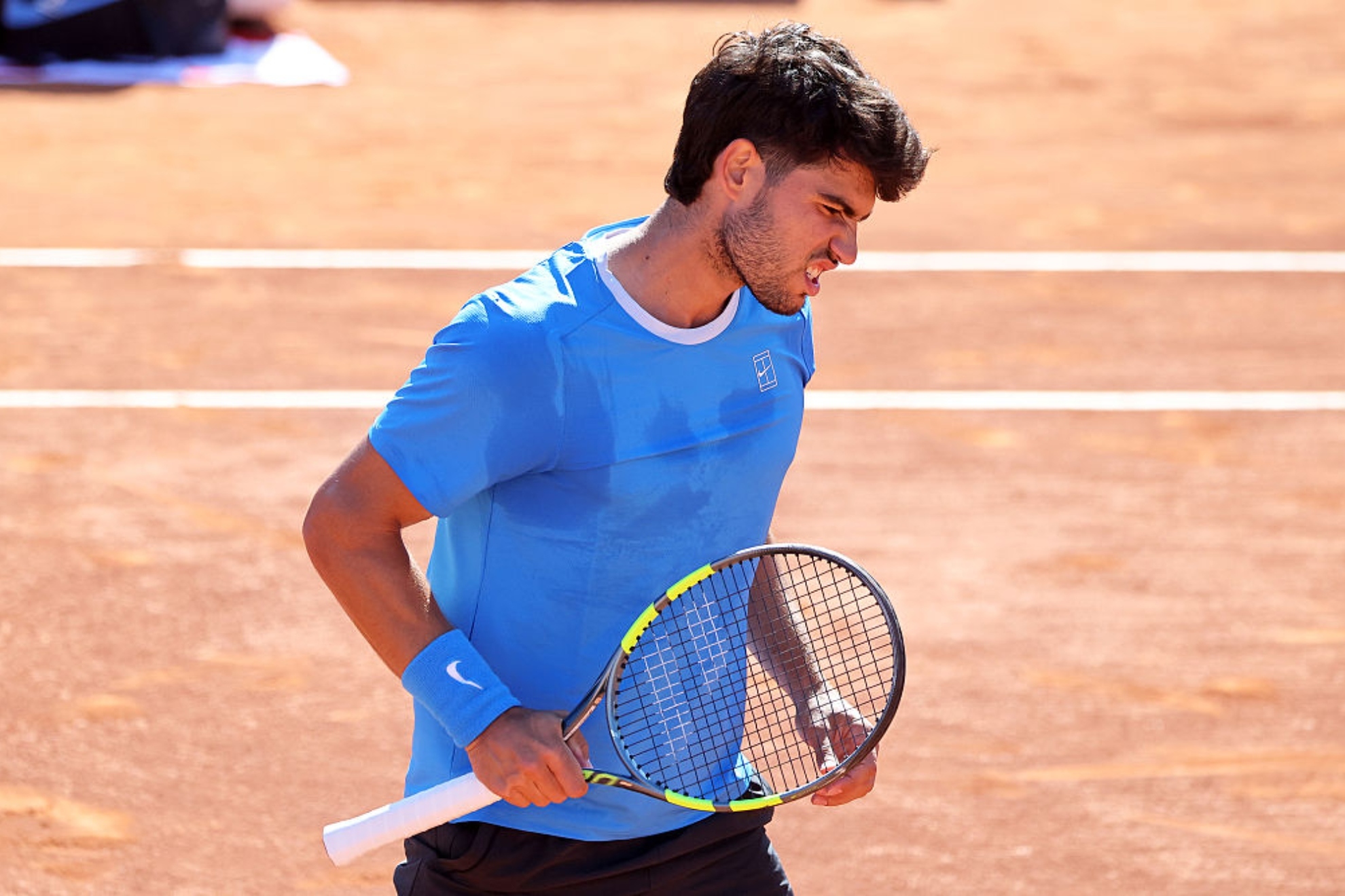 Un tenista joven viste camiseta azul y sostiene una raqueta de tenis en una cancha de tierra roja durante un partido.