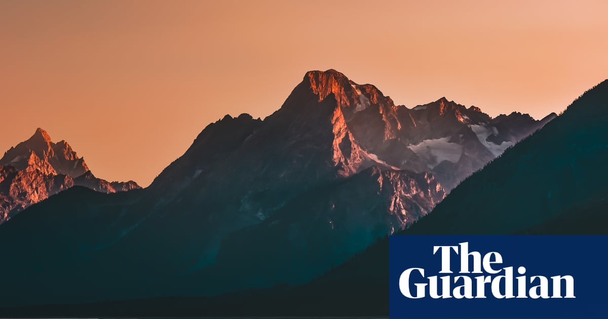 Rocky mountains with peaks illuminated by golden dawn light against a pale orange sky, with dark pine forests on the slopes.