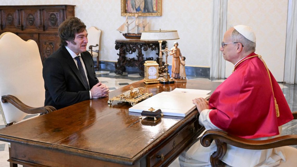 Un hombre de traje negro y corbata azul marino conversa con un prelado católico vistiendo sotana roja frente a un escritorio de madera en una sala de la Casa Rosada.