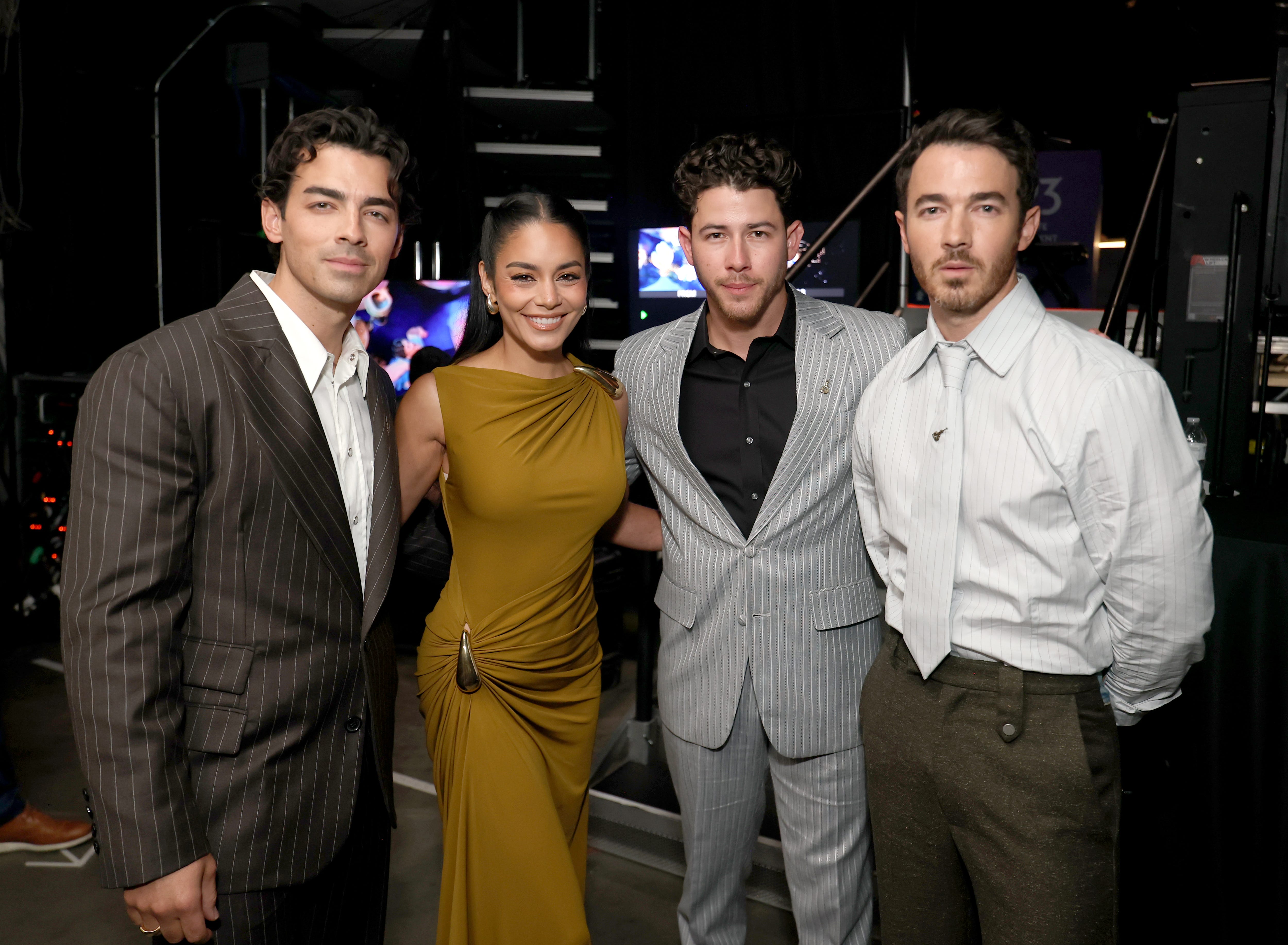 Four people stand together at an event, with two men in grey striped suits, a woman in a sleeveless gold dress and a man in a brown suit with white shirt, posing for a photograph on a dark stage with backlighting.