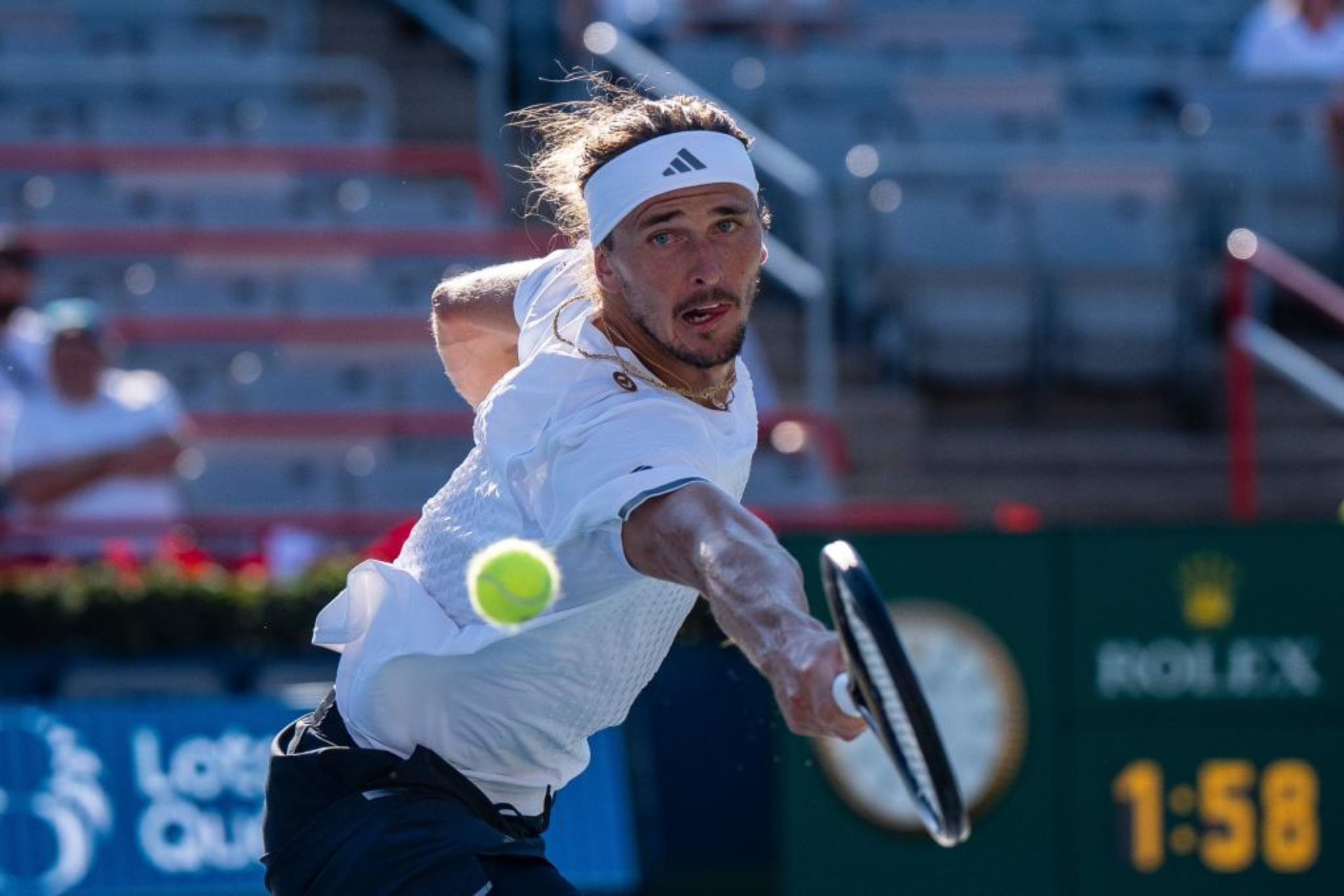 A male tennis player in white Adidas attire hits a forehand stroke during a professional tennis match on a hard court.