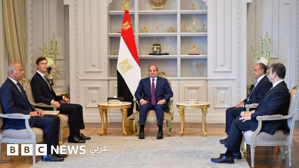 Un hombre en traje azul marino y corbata roja se sienta en el centro de una sala elegante con una bandera de Egipto detrás de él, rodeado de cuatro hombres en trajes oscuros sentados en sillas con marco dorado.