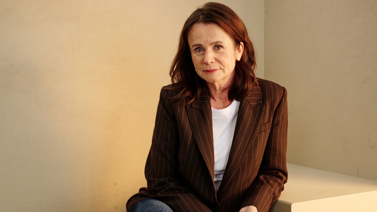 A woman with reddish-brown wavy hair wearing a brown pinstriped blazer over a white shirt sits against a neutral beige background.