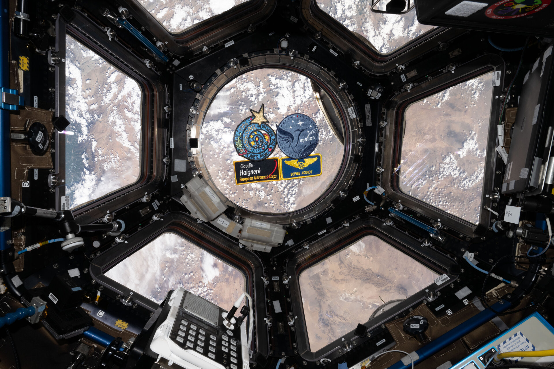 Vista desde la Cupola de la Estación Espacial Internacional mostrando la Tierra, con dos monedas conmemorativas y parches de misión de astronautas sostenidos frente a una ventana circular.
