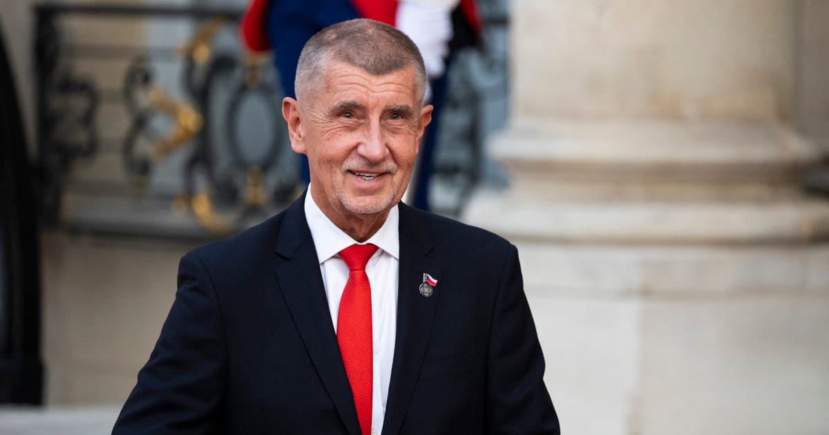 An elderly man in a dark suit and red tie poses for a photograph outside what appears to be an official building.