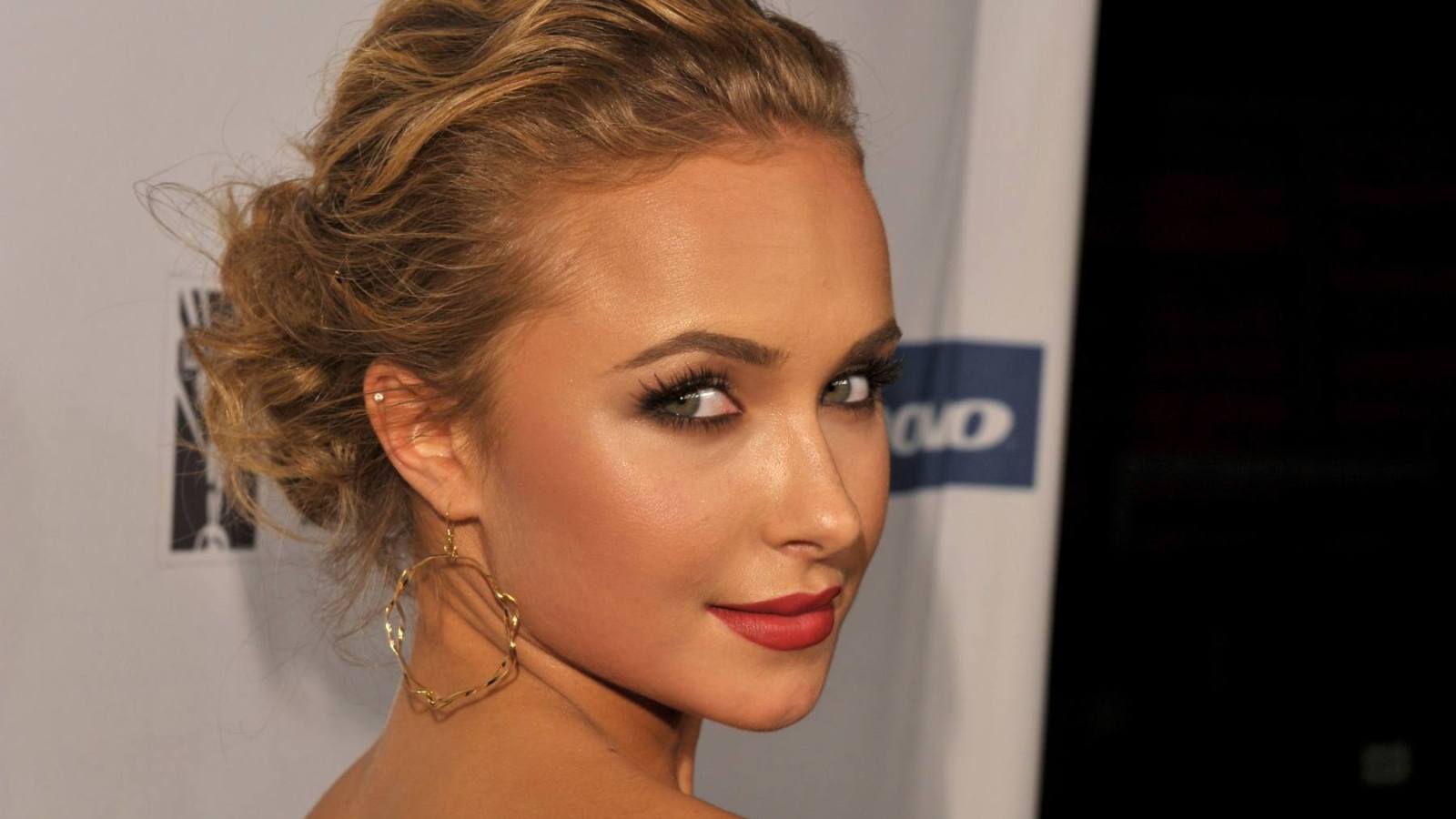 A woman with styled brown hair in an updo, wearing gold hoop earrings and red lipstick, photographed in profile at what appears to be an event with sponsor logos visible in the background.