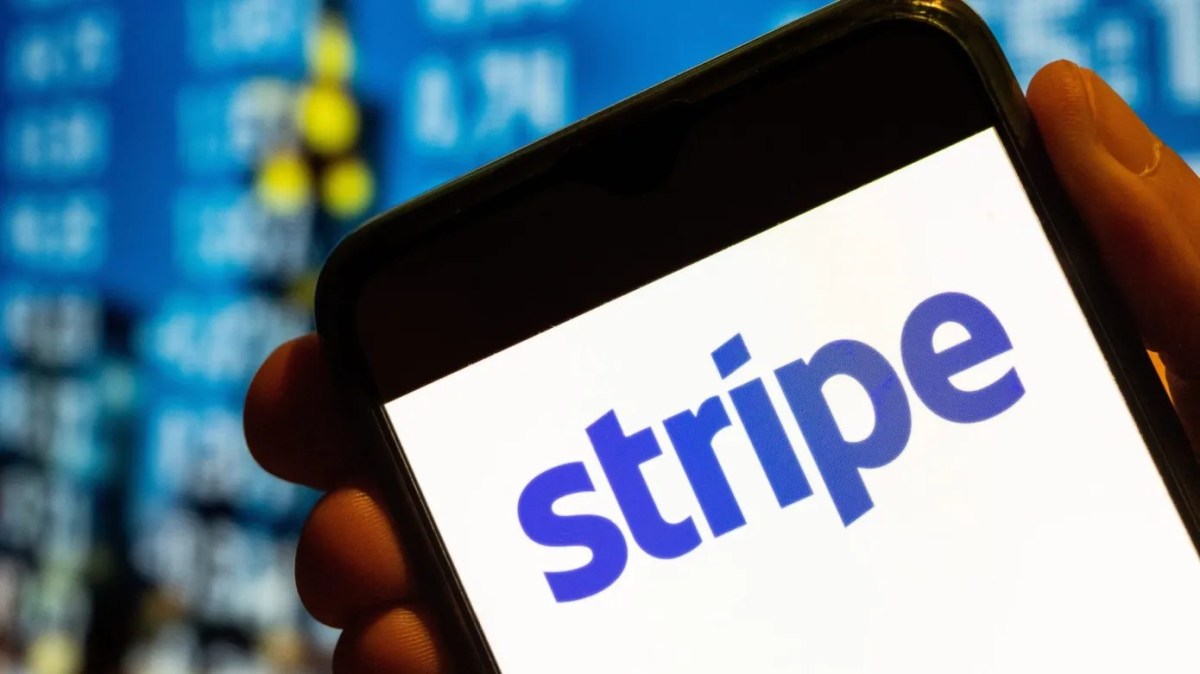 A hand holds a smartphone displaying Stripe's blue logo against a blurred background with data elements in blue and yellow tones.