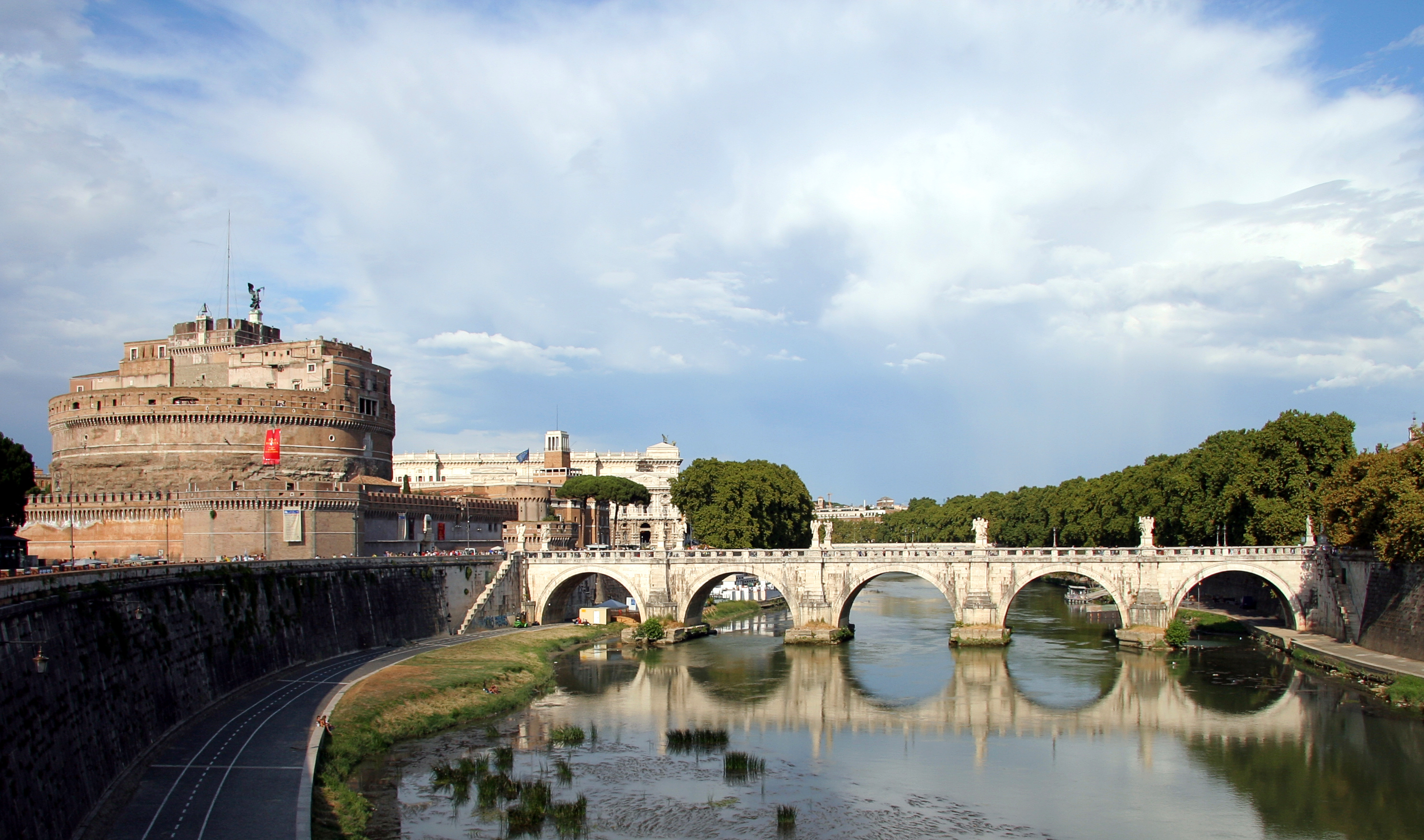 A historic castle tower and stone arch bridge span a river with green trees and buildings in the background under a partly cloudy sky.
