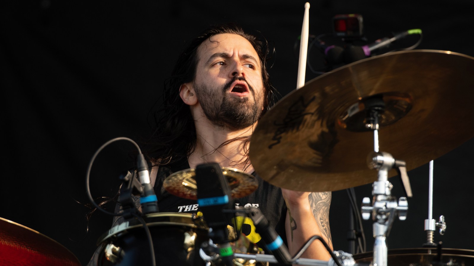 A long-haired man with a beard plays drums on stage, captured mid-performance with an expressive facial expression.