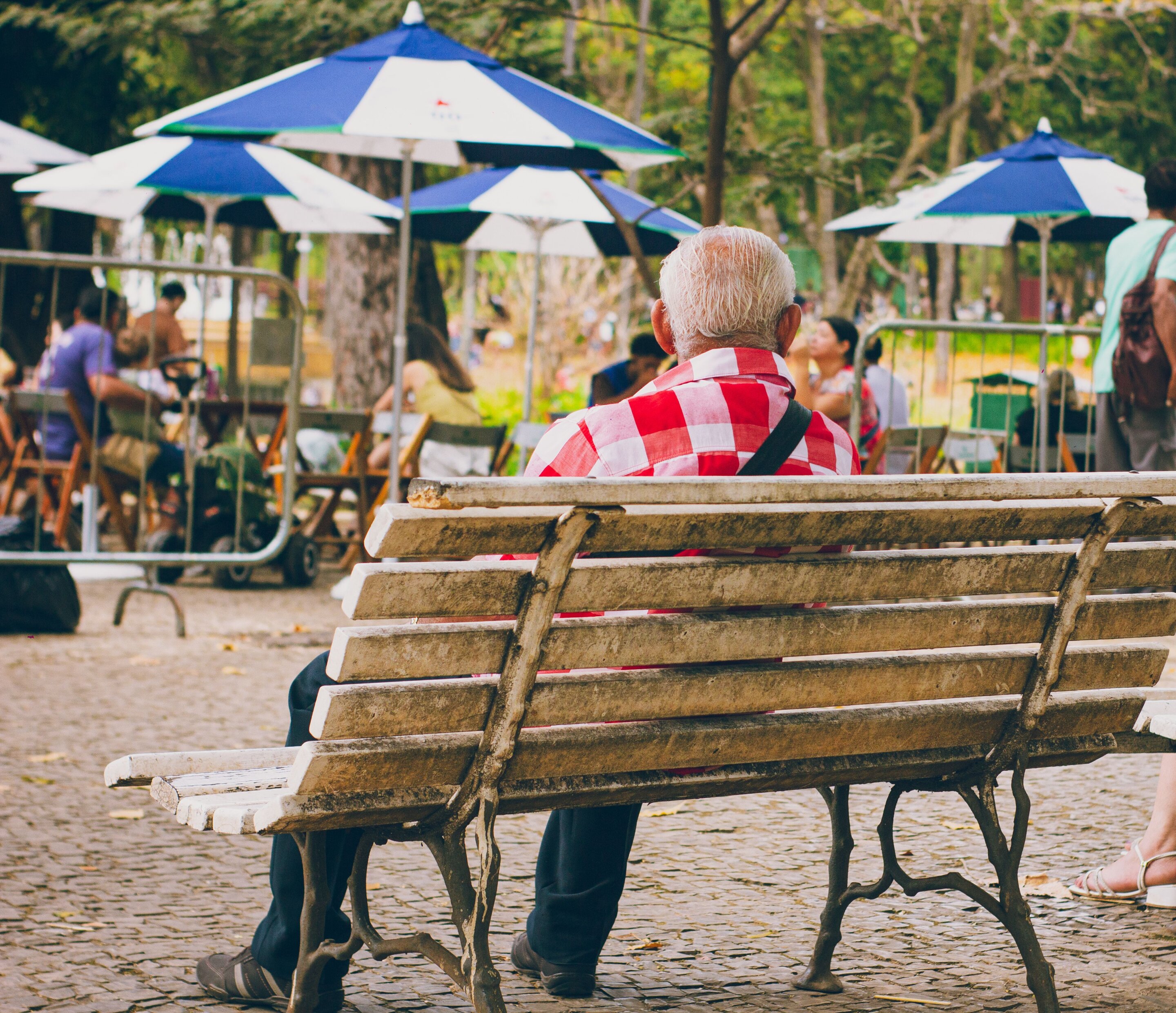 An elderly man with white hair wearing a red and white checkered shirt sits alone on a weathered wooden bench in a public plaza, looking toward a group of people gathered under blue and white umbrellas in the background.