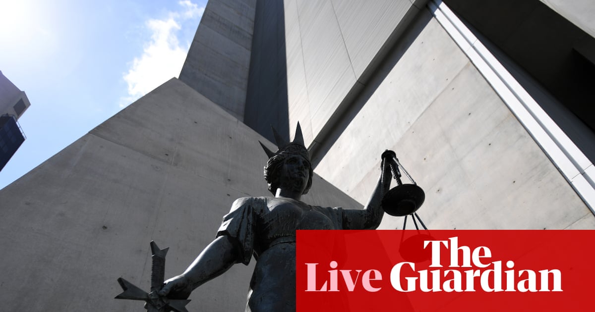 A bronze statue of Lady Justice holding scales and a sword, photographed from below against modern concrete government buildings and blue sky.