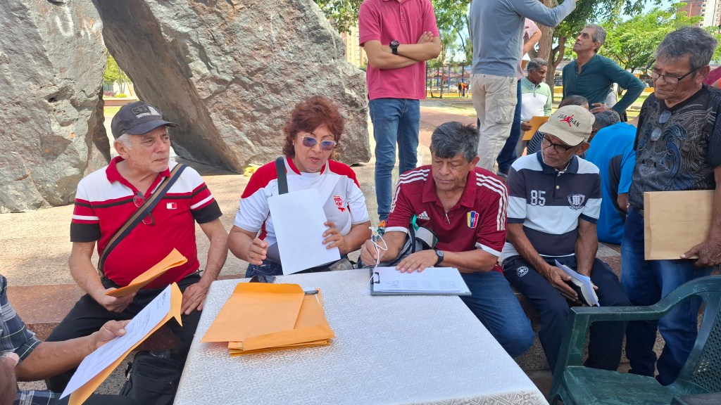 A group of retirees gather around a table in a park, with some signing documents while others observe.