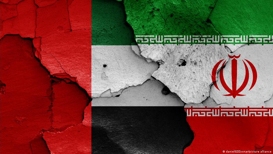 La bandera de Emiratos Árabes Unidos en rojo, blanco y negro aparece junto a la bandera de Irán en rojo, blanco y verde con el emblema del águila, ambas pintadas sobre una pared dañada y agrietada.