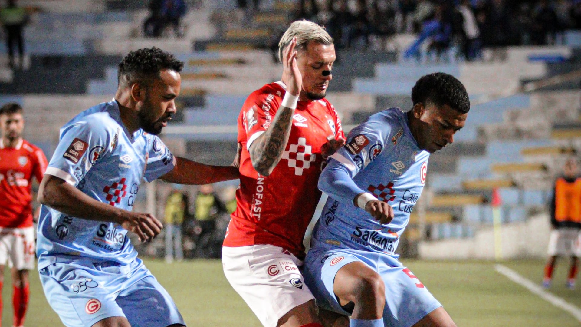 A player in red wearing number 9 disputes the ball with two players in blue during a football match in a stadium.
