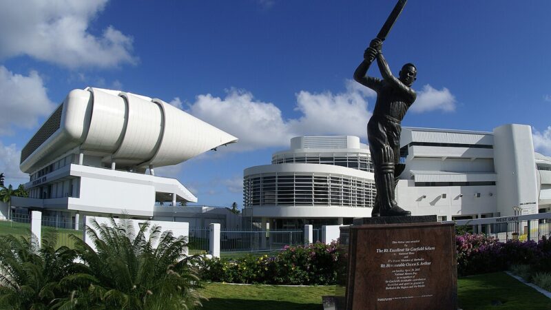 A bronze statue of a cricket player in batting position stands in front of a modern white building with a giant white sail and cylinder.
