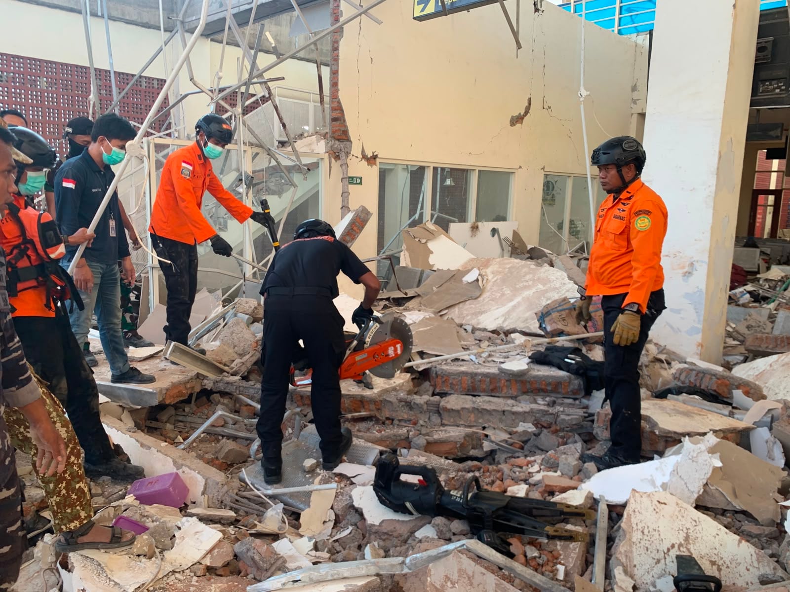 Rescatistas con chalecos naranja y negros trabajan entre escombros y estructuras dañadas de un edificio colapsado.