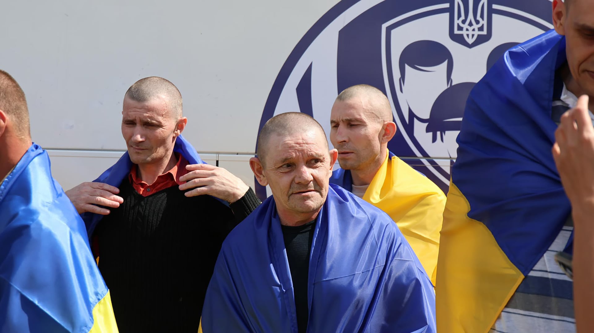 Several men wearing blue and yellow coats with the colours of the Ukrainian flag stand in front of a background with the coat of arms of Ukraine.