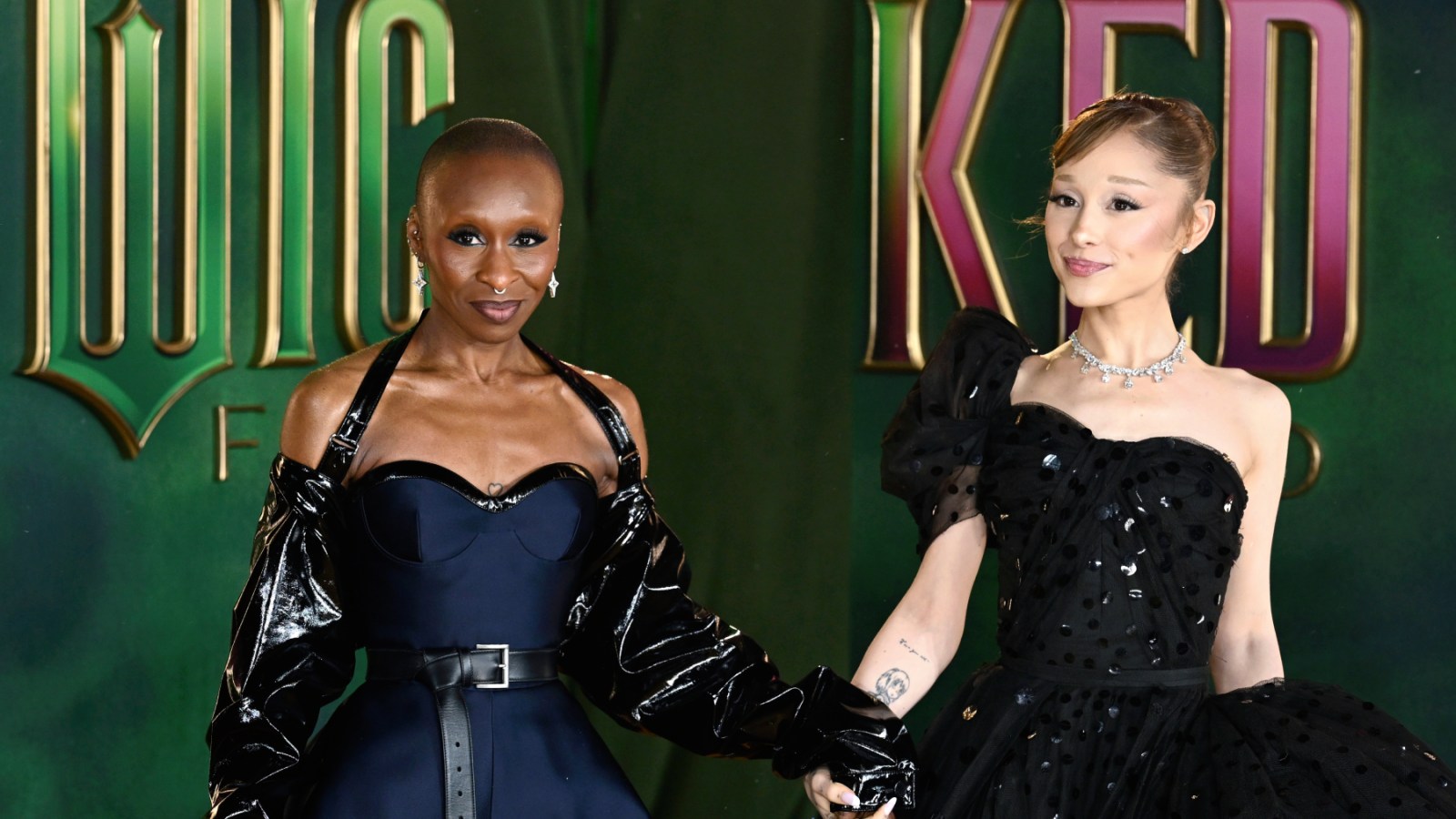 Two women in elegant black formal wear stand at a red carpet event in front of a green and purple branded backdrop.
