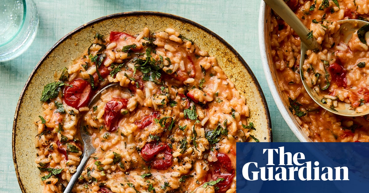 A plate of tomato risotto with fresh basil leaves, served on a cream-coloured ceramic plate with dark rim, accompanied by a glass of water and another portion in a white container in the background.