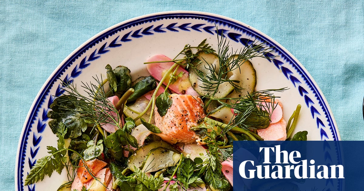 Un plato blanco y azul con decorados geométricos contiene ensalada de trucha confitada con calabacín a la parrilla, hierbas frescas, pétalos de rábano rosa y almendras, sobre un fondo de tela azul claro.