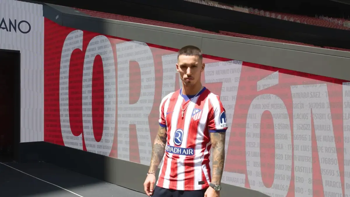 Un futbolista viste la camiseta roja y blanca del Atlético de Madrid y posa frente a un panel publicitario del estadio.