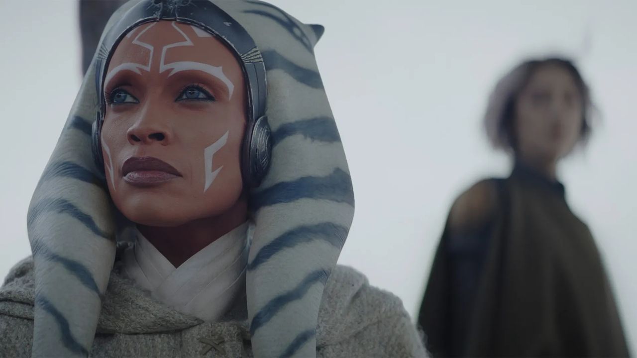A woman with white and red alien-style facial makeup, grey headdress with blue stripes and headphones, looking upward with a serious expression.