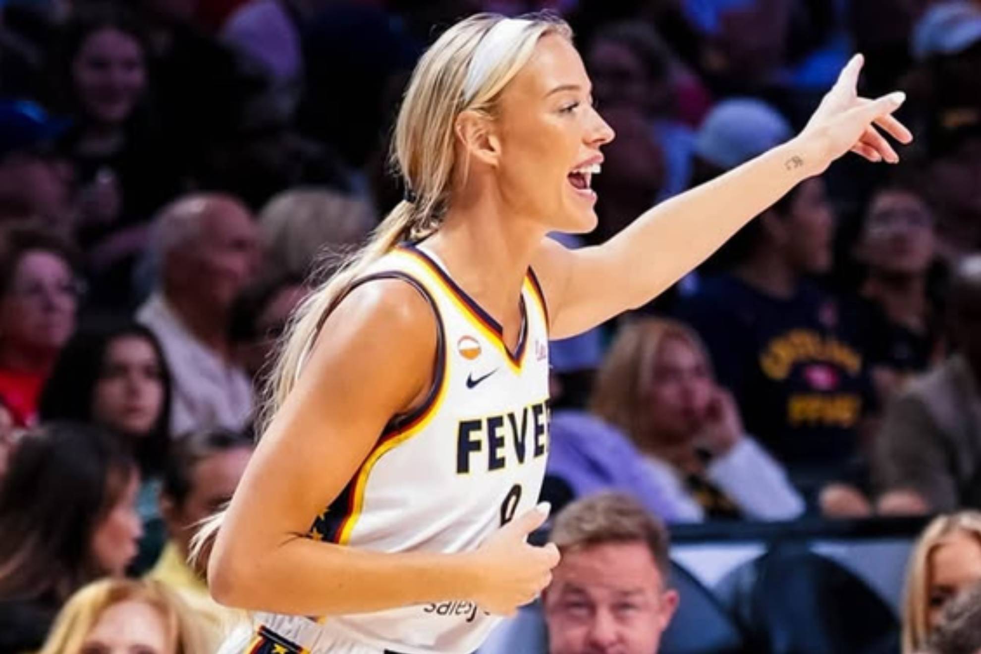 A blonde basketball player wearing a yellow and white Fever jersey with number 0 gestures with her arm extended while speaking on the court during a game with spectators in the background.
