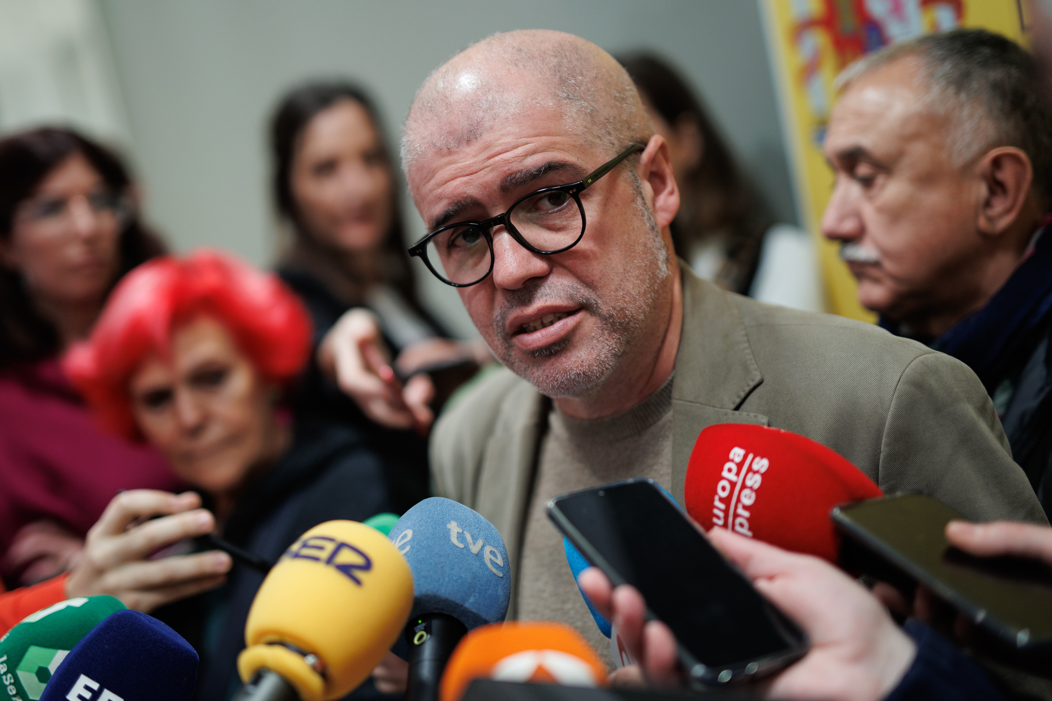 A man wearing glasses and a grey suit speaks into several media microphones, surrounded by journalists and people in the background.