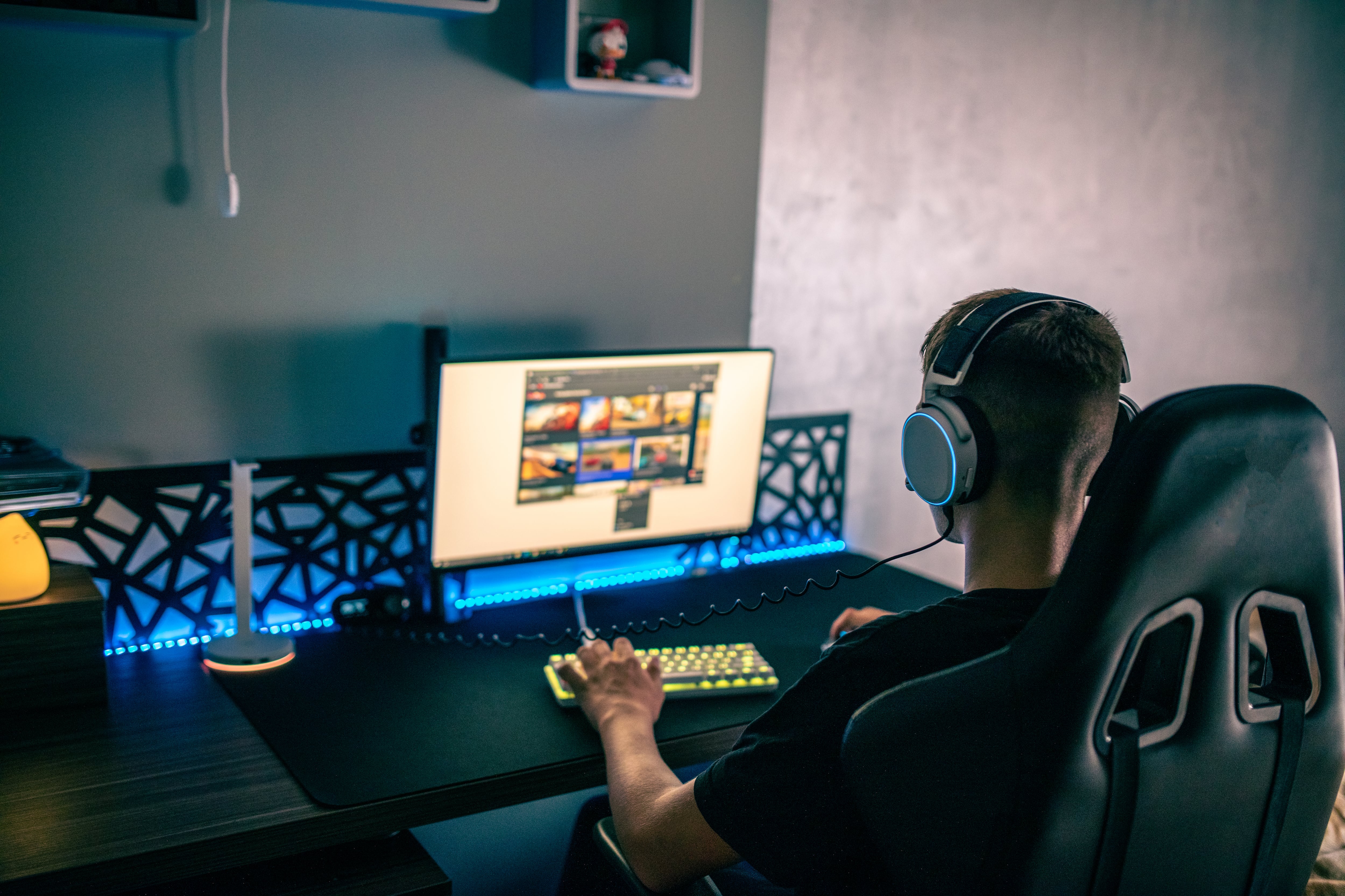 Un jugador de espaldas frente a un monitor de computadora, usando auriculares, con teclado iluminado en azul y amarillo en un escritorio gaming oscuro.